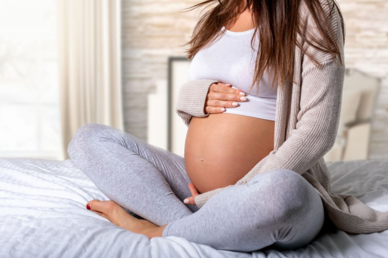Seção inferior de mulher grávida tocando a barriga enquanto está sentada na cama
