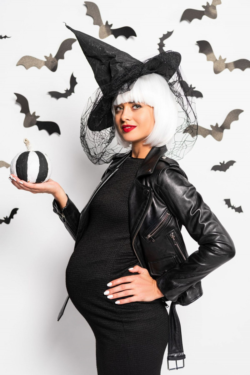 tehotná žena v čarodejníckom klobúku a parochni držiaca tekvicu na halloween