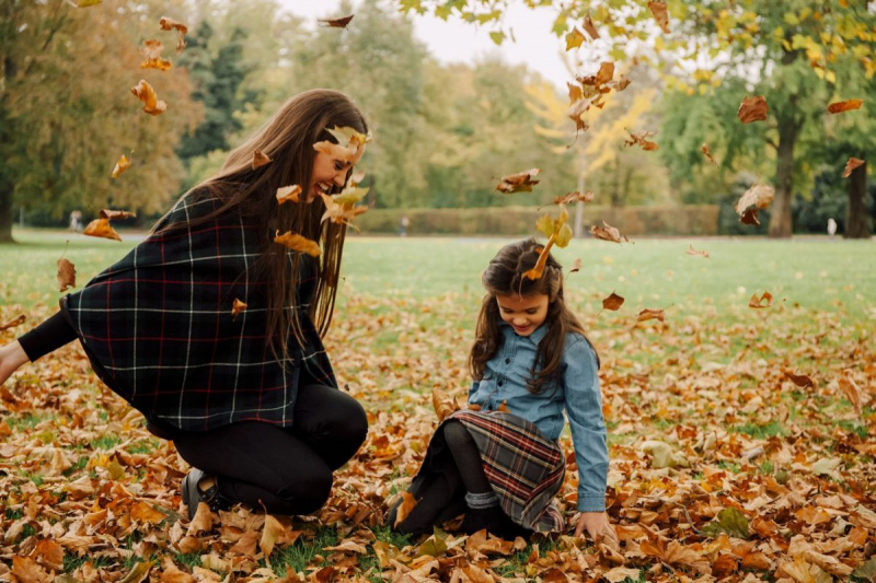 Млада жена и момиченце играят с есенни листа на поляна